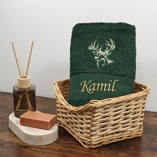 Dunkelgrünes Baumwollhandtuch in einem Weidenkorb, bestickt mit einer weißen Hirschkopf-Silhouette und dem Namen Kamil in goldener Schreibschrift, dekoriert mit Raumduft und Seife.