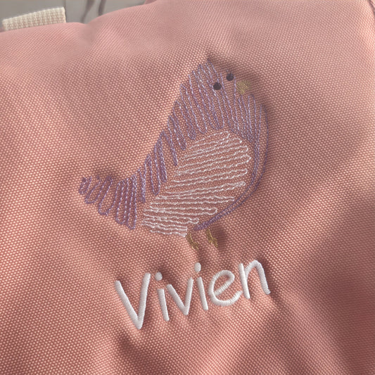 Nahaufnahme der präzisen Stickerei des lila Vogels und des Namens Vivien auf altrosafarbenem Rucksackstoff.