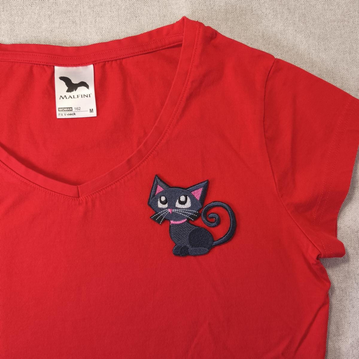 Rotes V-Ausschnitt-T-Shirt mit einem schwarzen Katzenaufnäher. Der Katzenaufnäher hat einen rosa Kragen und Schnurrhaare.