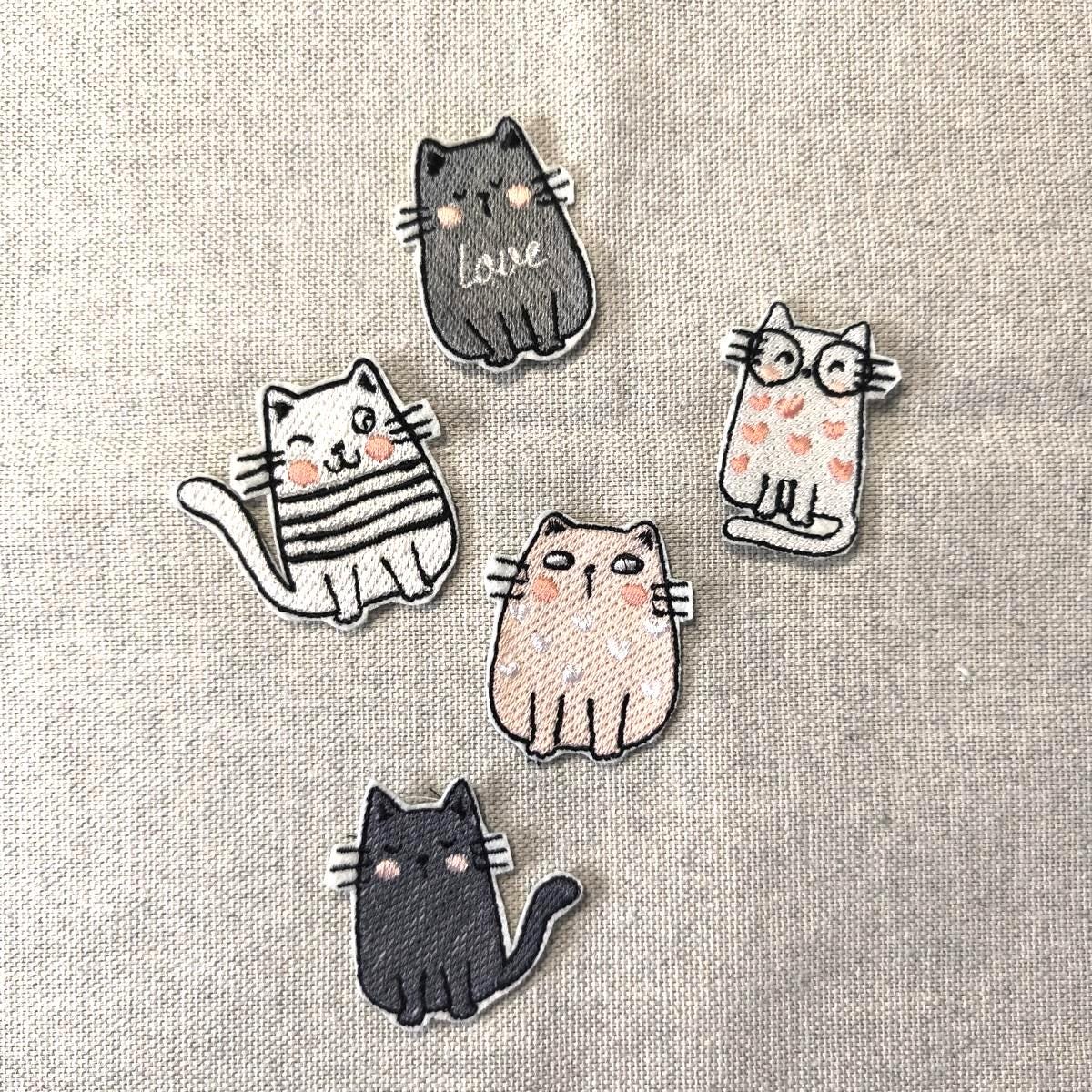Fünf verschiedene bestickte Katzen-Aufnäher (Patches) zum Aufbügeln, darunter eine Katze mit Brille, eine mit Love-Schriftzug und eine zwinkernde Katze, flach liegend auf Leinenstoff.