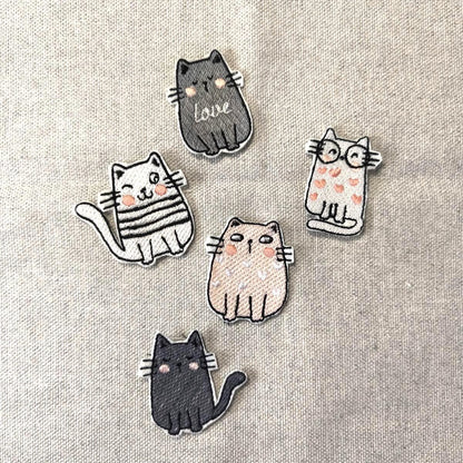 Fünf verschiedene bestickte Katzen-Aufnäher (Patches) zum Aufbügeln, darunter eine Katze mit Brille, eine mit Love-Schriftzug und eine zwinkernde Katze, flach liegend auf Leinenstoff.