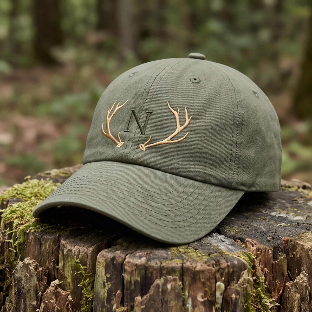Olivengrüne Baseball-Kappe aus Bio-Baumwolle mit gesticktem goldenen Hirschgeweih und Monogramm N auf einem Baumstamm im Wald.