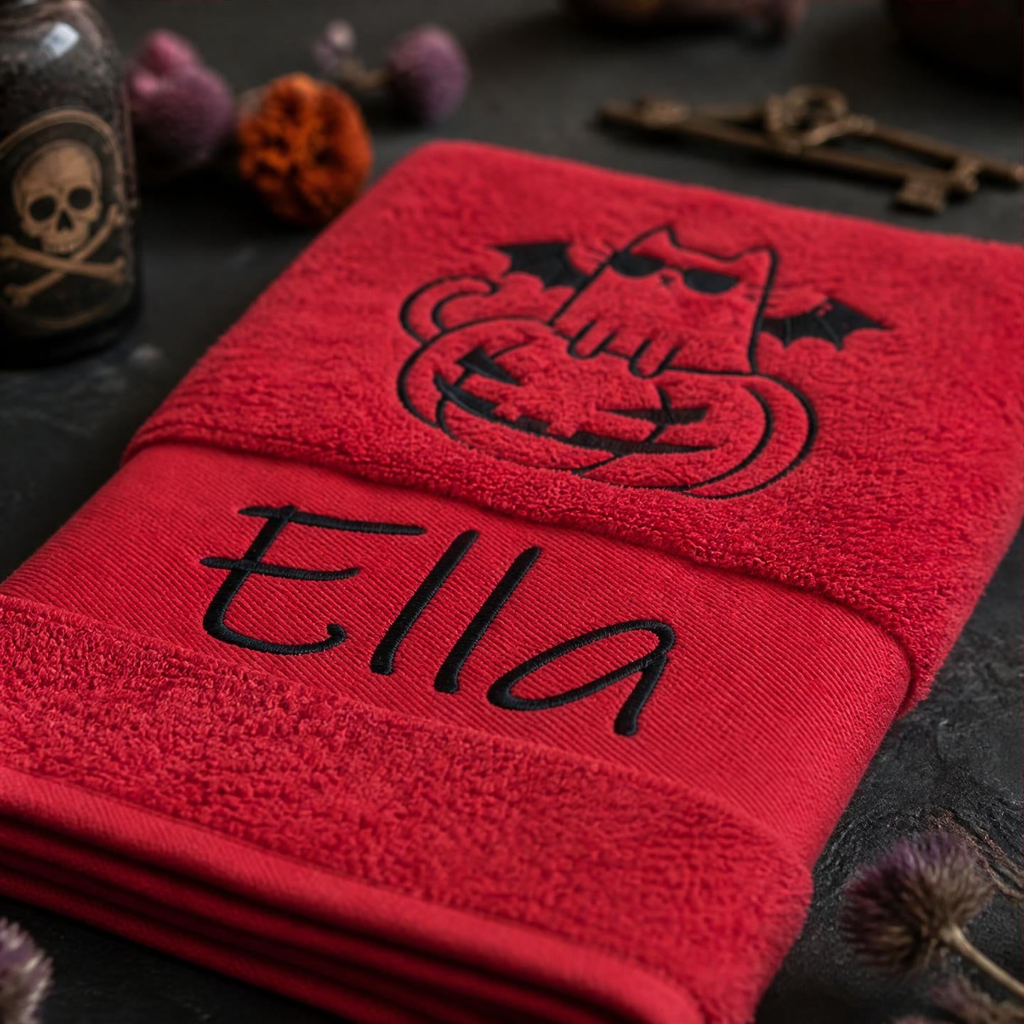 Gefaltetes rotes Handtuch mit schwarzer Namensbestickung Ella und Halloween-Katzen-Motiv neben Deko-Totenkopf