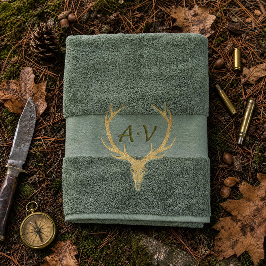 Olivgrünes handtuch mit goldener Hirschschädel und Geweih sowie Initialen Bestickung auf einem natürliche Waldboden Hintergrund.