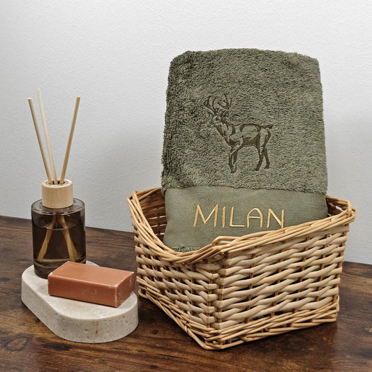 Olivgrünes besticktes Handtuch mit einem stolzen Hirsch-Motiv und dem Namen Milan in goldener Schrift, präsentiert in einem Weidenkorb mit Seife und Raumduft.