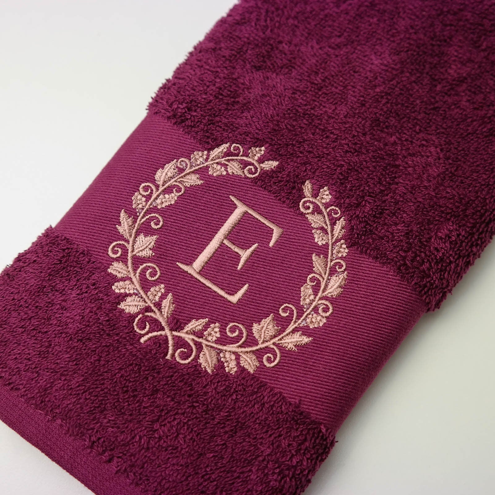 Berryfarbenes Badetuch mit einer rosa gestickten Weinblätterkranz und dem Buchstaben "E" in der Mitte