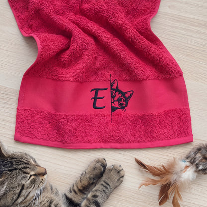 Ausgefaltetes pinkes Handtuch mit gesticktem Buchstaben E und schauender Katze neben schlafender getigerter Katze
