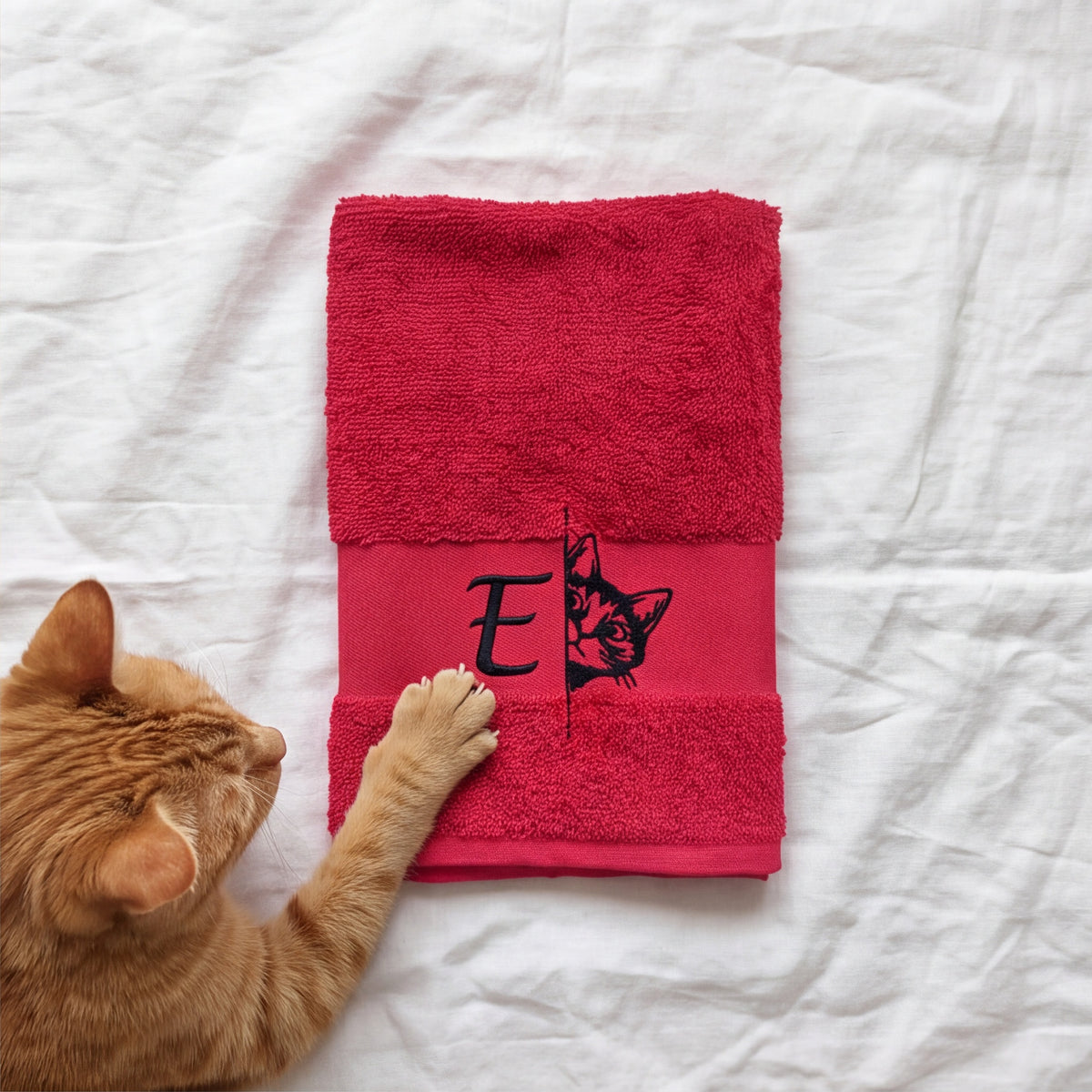 Gefaltetes pinkes Handtuch mit besticktem Monogramm E und Katze, berührt von einer orangen Katzenpfote auf weißem Linnen