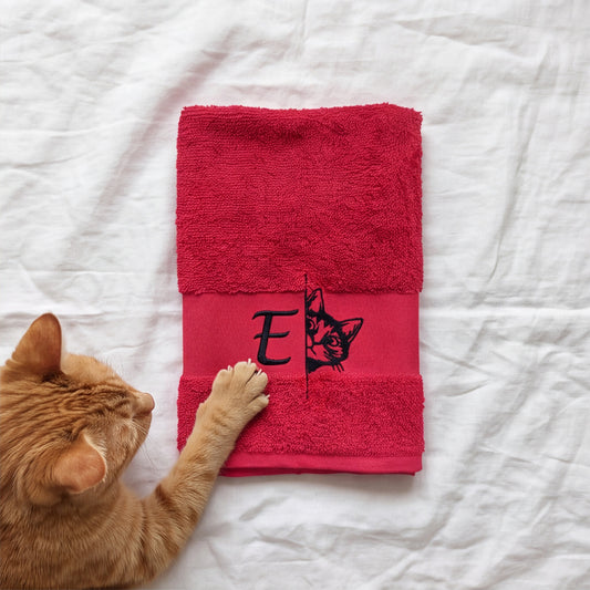 Gefaltetes pinkes Handtuch mit besticktem Monogramm E und Katze, berührt von einer orangen Katzenpfote auf weißem Linnen
