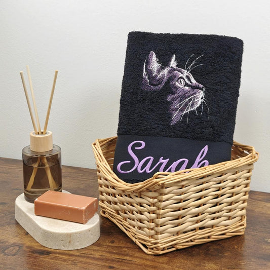 Schwarzes flauschiges Handtuch in einem Flechtkorb, bestickt mit einem lila-weißen Katzenkopf im Profil und dem Namen Sarah in fliederfarbener Schreibschrift, dekoriert mit Raumduft und Seife.