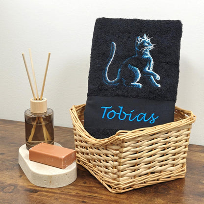 Schwarzes Duschtuch mit einer blau schimmernden Stickerei einer sitzenden Katze und dem Namen Tobias in blauer Schreibschrift, stilvoll arrangiert in einem hellen Flechtkorb.