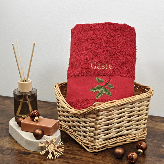 Rotes flauschiges Handtuch in einem Weidenkorb, bestickt mit einem grünen Stechpalmenzweig mit roten Beeren und dem Wort Gäste in goldener Schrift, weihnachtlich dekoriert mit braunen Kugeln und Strohsternen.