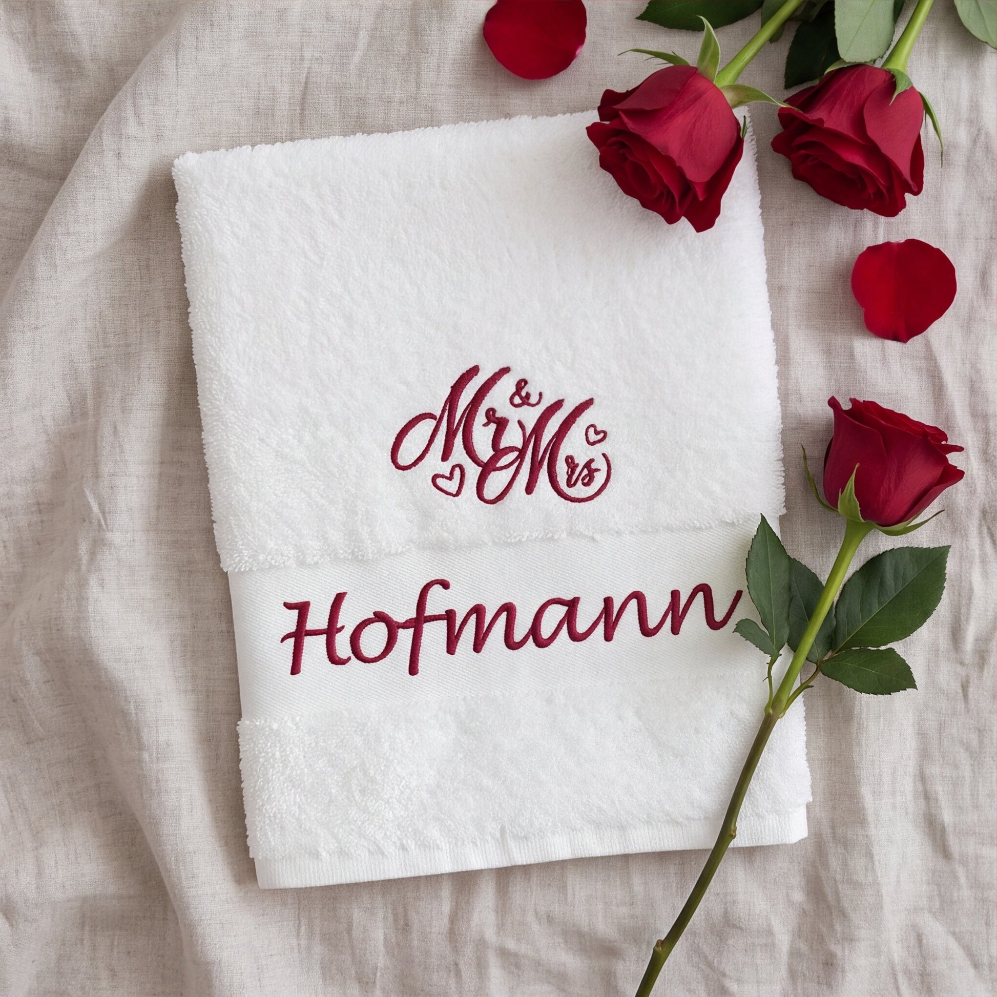 Gefaltetes weißes Handtuch mit besticktem Namen Hofmann und Mr & Mrs Motiv auf beigefarbenem Leinen mit roter Rose