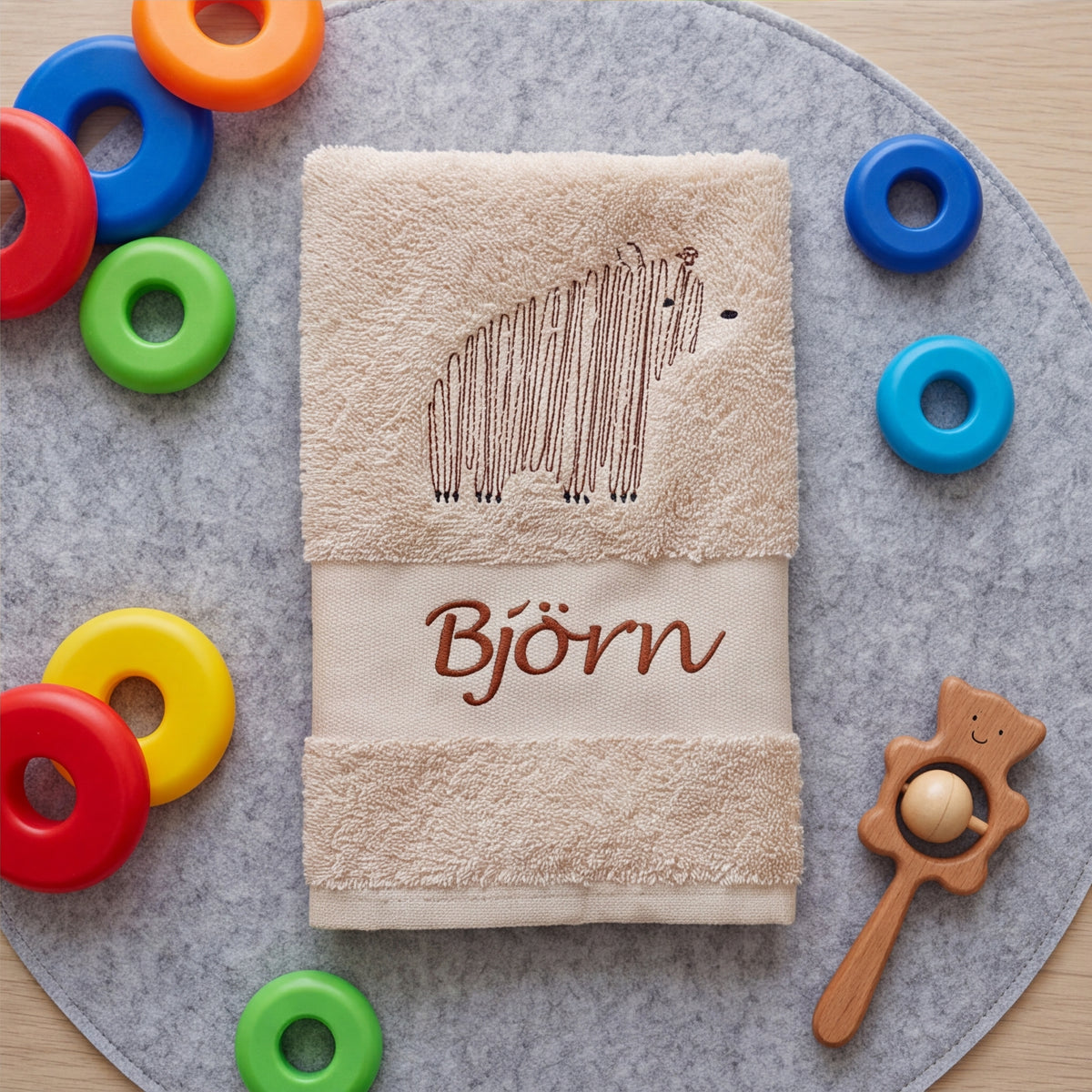 Beige zusammengelegtes Kinderhandtuch mit Bären-Stickerei und Namenszug Björn, umgeben von bunten Stapelringen auf einer grauen Filzunterlage.