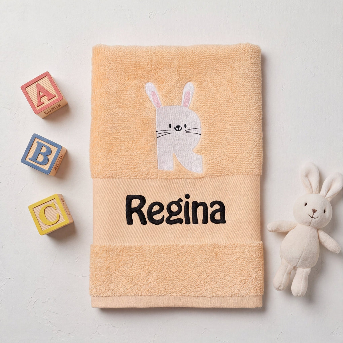 Personalisiertes Babyhandtuch mit Hasen-Buchstabe R und dem Namen Regina neben bunten ABC-Holzbausteinen und einem Kuschelhasen.