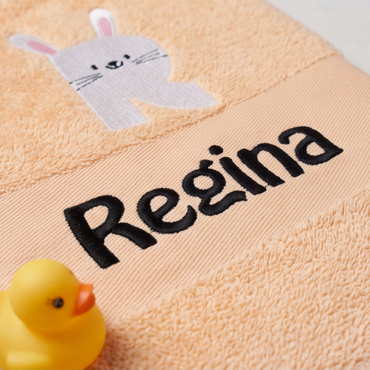 Nahaufnahme der schwarzen Namensstickerei Regina und der präzisen Hasen-Initialen R auf flauschigem Baumwoll-Frotteestoff neben einer gelben Badeente.