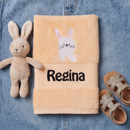 Pfirsichfarbenes Kinderhandtuch mit gesticktem Hasen-Initial R und Namenszug Regina auf Jeansstoff neben einem Kuschelhasen und Kindersandalen.