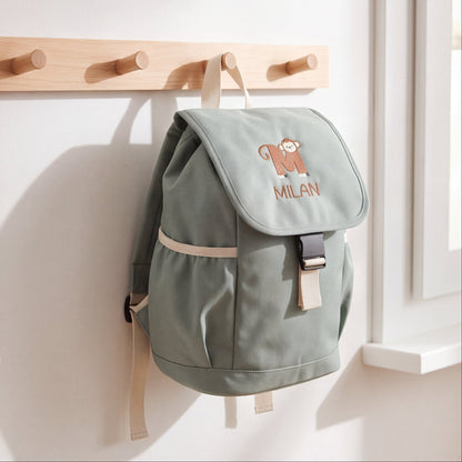 Personalisierter Kinderrucksack in Dusty Green mit Namensstickerei Milan und Tier-Buchstabe M an einer Holz-Garderobenleiste.
