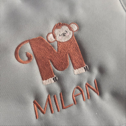Nahaufnahme der präzisen Stickerei des Affen-Motive-Initialen M und des Namens Milan auf robustem Rucksackstoff.