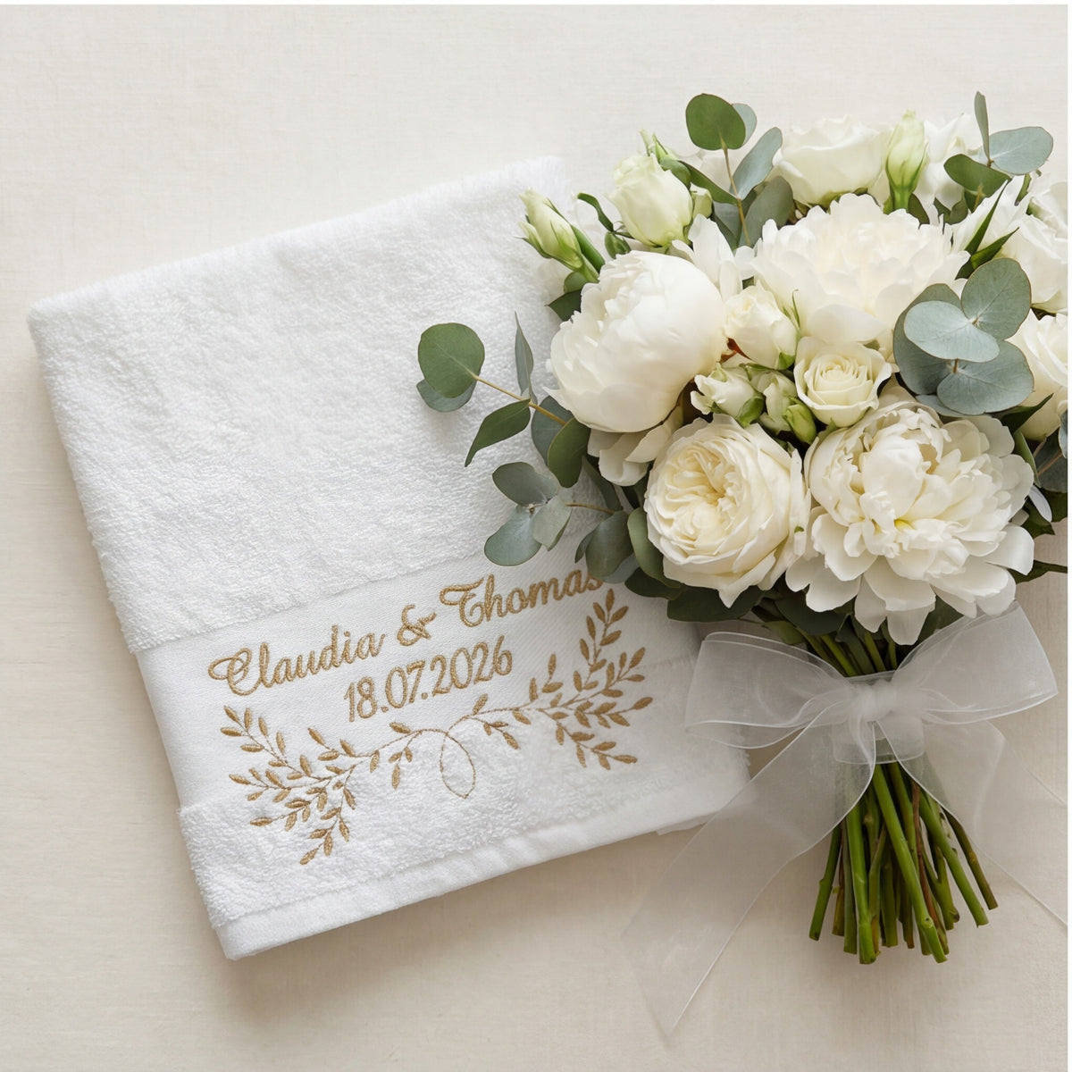 Weißes Handtuch mit goldener Namen, Datum und Blumenstickerei umgeben von Hochzeitsdekoration.