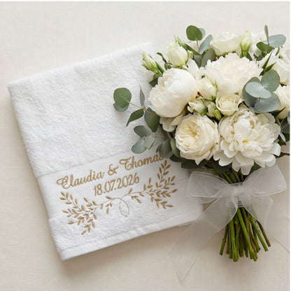 Weißes Handtuch mit goldener Namen, Datum und Blumenstickerei umgeben von Hochzeitsdekoration.