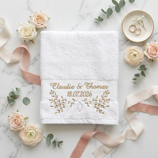 Weißes Handtuch mit goldener Namen, Datum und Blumenstickerei umgeben von Hochzeitsdekoration.