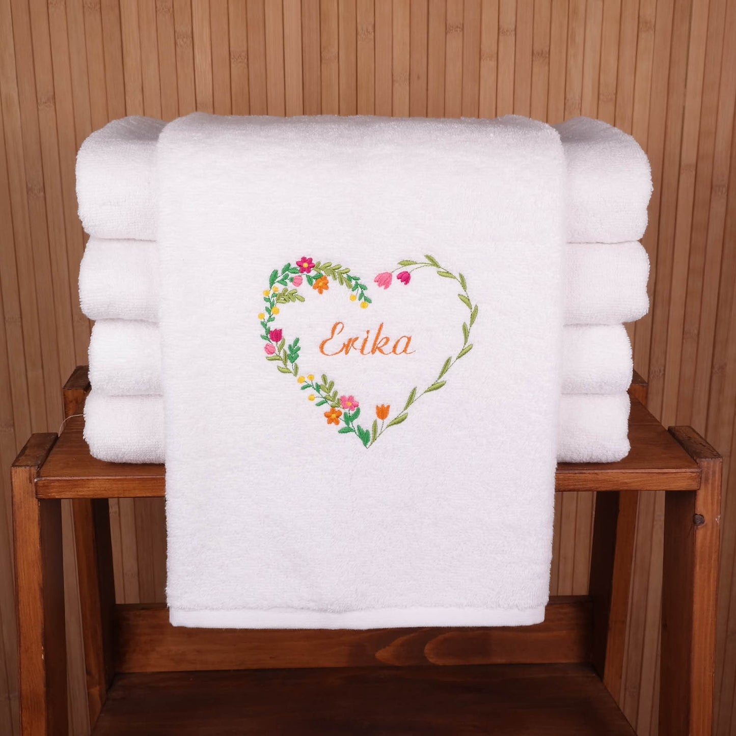 Weißes flauschiges Handtuch, bestickt mit einem filigranen Herz aus bunten Frühlingsblumen und dem Namen Erika in oranger Schreibschrift, präsentiert auf einem rustikalen Holzregal.