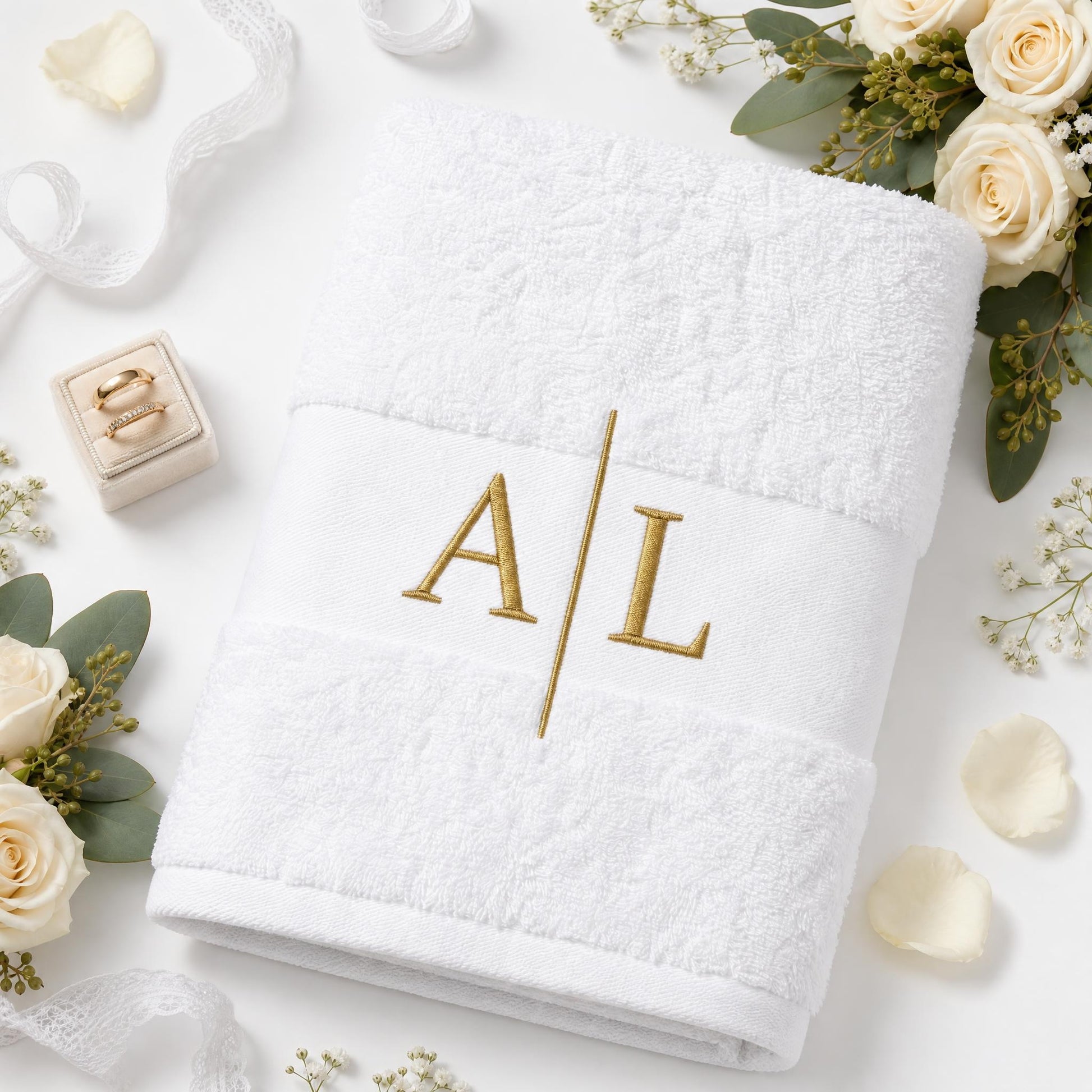 Ein weißes Hochzeitshandtuch mit goldenen Initialen und einer Trennlinie bestickt etwas seitlich liegend. Rundherum liegen Hochzeitsblumen, Dekoration sowie Verlobungsringe.