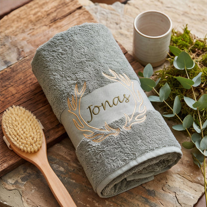 Aufgerolltes olives Frottee-Handtuch mit goldenem Geweih-Stick und Namenszug Jonas auf Holzplatte mit Wellness-Massagebürste und Eukalyptus.