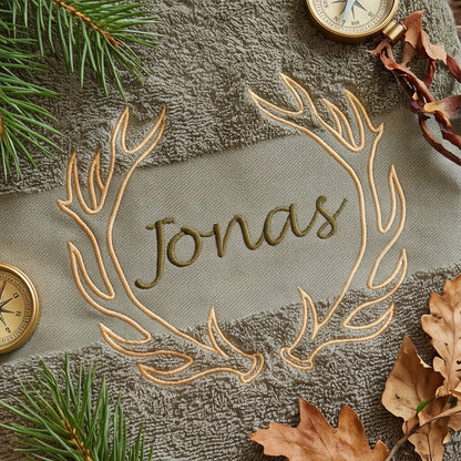 Nahaufnahme der goldenen Geweih-Stickerei und der dunkelgrünen Namensstickerei Jonas auf olivem Baumwollstoff umgeben von Tannenästchen und Kompass.